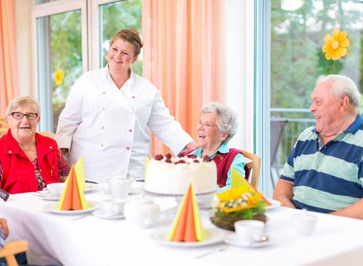 Küchenmitarbeiterin steht am Tisch mit drei Bewohnern und hat eine Torte serviert
