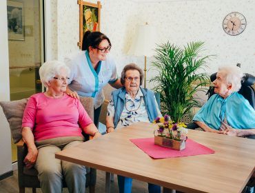 Bewohnerinnen sitzen zusammen am Tisch und werden von Pflegerin betreut
